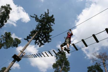 Eine Person mit einem Klettergurt überquert eine Hochseilgartenbrücke, die zwischen Bäumen vor einem blauen Himmel mit vereinzelten Wolken hängt.