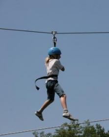 Eine Person, die einen blauen Helm und ein Gurtzeug trägt, fährt auf einer Seilrutsche gegen einen klaren Himmel.