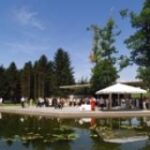 An einem sonnigen Tag versammeln sich Menschen unter Zelten an einem Teich mit Seerosenblättern, umgeben von Bäumen und Grasflächen.