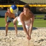Zwei Männer spielen Beachvolleyball; ein Spieler taucht, um den Ball zu schlagen, während der andere hinter ihm auf einem Sandplatz steht.
