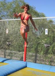 Eine Frau im roten Bikini springt in der Luft auf einem Trampolin im Freien, mit einem Sicherheitsnetz und Grünpflanzen im Hintergrund.
