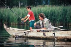 Ein Mann und ein Junge angeln von einem kleinen Holzboot aus auf einem ruhigen See, mit hohem Schilf im Hintergrund.