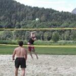 Menschen spielen Beachvolleyball im Freien auf einem Sandplatz mit Bäumen und Bergen im Hintergrund.