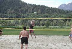 Menschen spielen Beachvolleyball im Freien auf einem Sandplatz mit Bäumen und Bergen im Hintergrund.