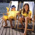 Zwei Kinder sitzen an einem sonnigen Tag auf Liegestühlen auf einem hölzernen Steg an einem See, lächeln und halten Eistüten in der Hand.