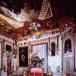 Verziertes Zimmer im Barockstil mit kunstvollen Deckenfresken, goldverzierten Möbeln und einem zentralen Bett in Rosa und Gold unter einer reich verzierten Decke.