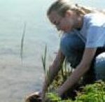 Eine Person mit langen blonden Haaren, die zu einem Zopf geflochten sind, hockt am Rande eines Teiches und streckt ihre Hände ins Wasser.