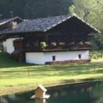 Ein rustikales Holzhaus mit Blumenkästen auf dem Balkon steht in der Nähe eines kleinen Teichs mit Zaun, umgeben von Gras und Bäumen.