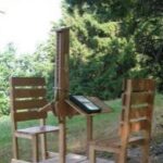 Zwei Holzstühle und ein kleiner Tisch sind im Freien aufgestellt; der Tisch hat einen ungewöhnlichen vertikalen Aufsatz, der ein Buch oder ein Tablet hält. Im Hintergrund sind Bäume und Grünflächen zu sehen.