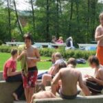 Mehrere hemdsärmelige Jungen versammeln sich im Freien in der Nähe eines Pools, einige sitzen auf Betonstufen, andere stehen, mit Bäumen und weiteren Menschen im Hintergrund.