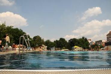 Freibad mit Menschen, die sich entspannen und schwimmen, umgeben von Bäumen und Gebäuden unter einem teilweise bewölkten Himmel.