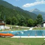 Freibad, umgeben von Gras, Bäumen und Bergen unter einem teilweise bewölkten Himmel, mit mehreren Gebäuden in der Nähe und einer Rutsche, die in den Pool führt.