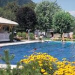 Die Menschen schwimmen und entspannen sich an sonnigen Tagen in einem von Bäumen, Blumen und einem Holzpavillon umgebenen Freibad.