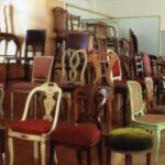 Eine Ausstellung verschiedener antiker Holzstühle mit gepolsterten Sitzen, die in Reihen auf gestuften Holzplattformen in einem Museum oder einer Galerie angeordnet sind.