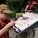 Ein Junge mit hellbrauner Mütze interagiert mit einem Saturn-Informationsdisplay im Freien; im Hintergrund stehen zwei Mädchen, von denen eines eine rote Tupfenmütze trägt.