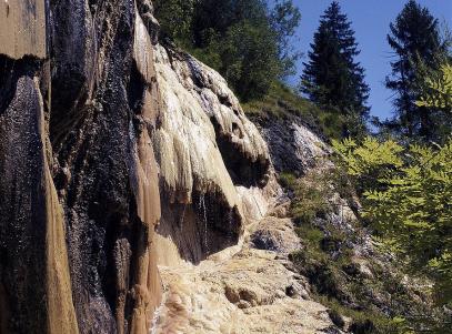 Felsen mit mineralischen Ablagerungen und spärlicher Vegetation, mit Bäumen und blauem Himmel im Hintergrund.