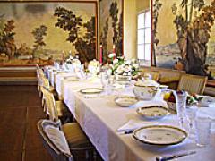 Ein langer, eleganter Esstisch, gedeckt mit feinem Porzellan und floralen Tafelaufsätzen in einem Raum mit malerischen Wandbildern und natürlichem Licht durch ein Fenster.