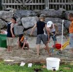 Fünf Kinder spielen zusammen in einem Sandgebiet mit Spielzeug und einer Rutsche, mit großen Felsen und einem Zaun im Hintergrund.