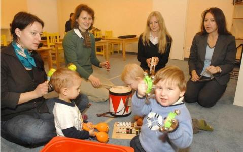 In einem hell erleuchteten Klassenzimmer sitzen drei Erwachsene mit drei kleinen Kindern auf dem Boden, die alle Musikinstrumente wie Maracas und Trommeln in der Hand halten.
