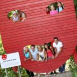 Eine Gruppe von Menschen posiert durch ovale Ausschnitte in einer großen roten Struktur in Form eines lächelnden Gesichts, mit Bäumen im Hintergrund.