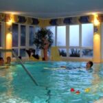 Mehrere Personen schwimmen und entspannen sich in einem Hallenbad mit großen Fenstern und gelben Wänden; Poolspielzeug schwimmt auf dem Wasser.