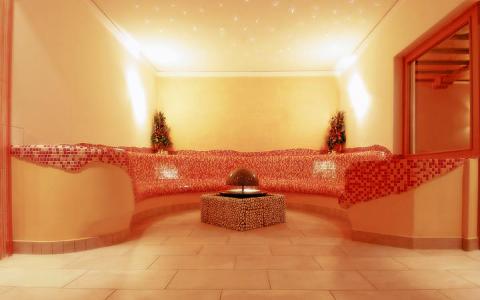 Ein sanft beleuchteter Raum mit einer geschwungenen Mosaikbank, zwei Topfpflanzen und einem zentralen Brunnen auf einem gefliesten Boden.