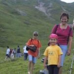 Eine Gruppe von Menschen, darunter Kinder und Erwachsene, geht auf einem grasbewachsenen Hügel mit einem Berg im Hintergrund spazieren.