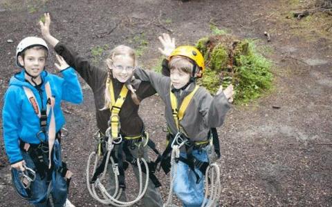 Drei Kinder mit Helmen und Klettergurten stehen im Freien auf der Erde, lächeln und heben die Arme.