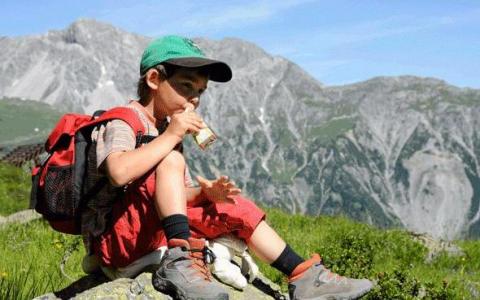 Ein Junge mit grüner Mütze und roter Hose sitzt auf einem Felsen und isst eine Kleinigkeit, während im Hintergrund die Berge zu sehen sind.