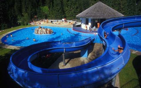 Außenschwimmbad mit blauer Wasserrutsche, umgeben von Bäumen. Mehrere Leute schwimmen und spielen im Pool und faulenzen in der Nähe.