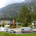 Mehrere Wohnmobile und Autos sind in der Nähe eines beflaggten Gebäudes vor der Kulisse bewaldeter Berge geparkt.