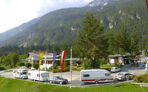 Mehrere Wohnmobile und Autos sind in der Nähe eines beflaggten Gebäudes vor der Kulisse bewaldeter Berge geparkt.