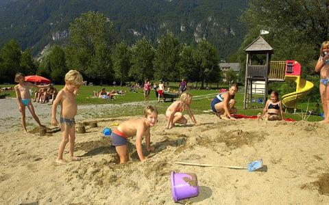 Kinder spielen im Sand auf einem Spielplatz mit Rutsche und Klettergerüst, während sich Erwachsene und andere Kinder im Hintergrund auf einer Wiese entspannen und spielen.