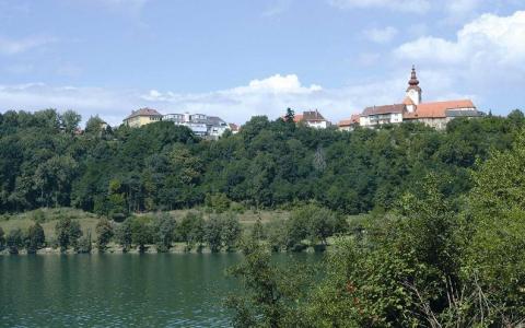 Ein kleines Dorf mit einer Kirche und mehreren Gebäuden liegt auf einem baumbestandenen Hügel und blickt auf ein ruhiges Gewässer.