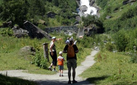 Eine Familie mit zwei Kindern geht auf einem Weg durch grüne Hügel in Richtung einer Steinbrücke und eines Wasserfalls im Hintergrund.