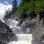 Wasser stürzt in Kaskaden einen felsigen Wasserfall hinunter, umgeben von grünen Bäumen und Sträuchern unter einem teilweise bewölkten Himmel.