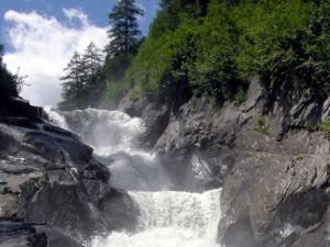 Wasser stürzt in Kaskaden einen felsigen Wasserfall hinunter, umgeben von grünen Bäumen und Sträuchern unter einem teilweise bewölkten Himmel.