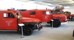 In einem Museum sind mehrere alte rote Feuerwehrautos ausgestellt, vor denen jeweils Schilder mit einer Beschreibung der Fahrzeuge angebracht sind.
