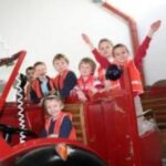 Eine Gruppe von Kindern mit orangefarbenen Sicherheitswesten sitzt und steht auf einem alten roten Feuerwehrauto in einem Gebäude.