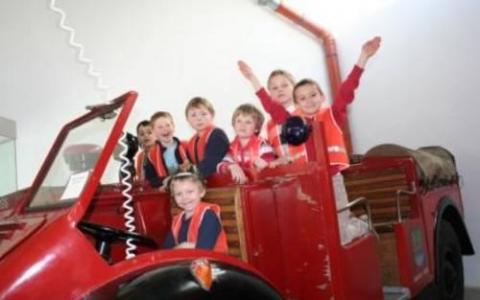 Eine Gruppe von Kindern mit orangefarbenen Sicherheitswesten sitzt und steht auf einem alten roten Feuerwehrauto in einem Gebäude.