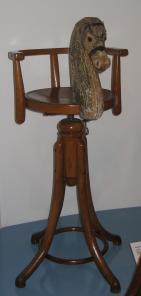 Ein hölzerner Drehstuhl mit einem geschnitzten Tierkopf, möglicherweise ein Pferd, der an der Rückenlehne befestigt ist.