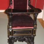 Verschnörkelter antiker Holzstuhl mit hoher Rückenlehne, rotem Samtbezug, detaillierten Schnitzereien, schwarzer Lackierung und Krallenfüßen, ausgestellt in einer Museumsumgebung.