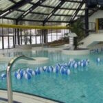 Hallenbad mit vielen blauen und weißen Strandbällen, die auf dem Wasser schwimmen, umgeben von Glaswänden und einem Glasdach.