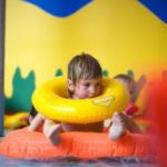 Ein Kind in einem Pool trägt einen gelben Schwimmring um den Hals und liegt auf einem orangefarbenen Schwimmkörper. Im Hintergrund sind bunte, abstrakte Formen zu sehen.