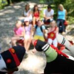 Im Vordergrund sind zwei Sockenpuppen zu sehen, während im Hintergrund eine Gruppe von sieben Personen im Freien sitzt und unscharf ist.