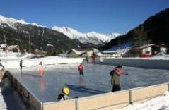 Menschen laufen Schlittschuh auf einer schneebedeckten Eisbahn im Freien, im Hintergrund sind Berge und Häuser unter einem klaren blauen Himmel zu sehen.