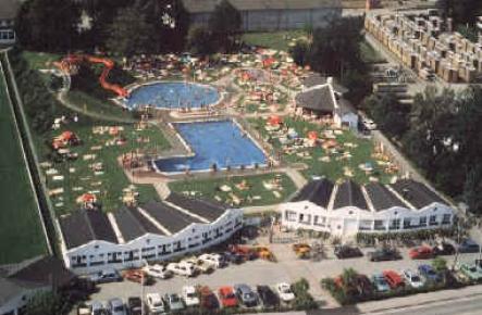 Luftaufnahme eines Freibadkomplexes mit zwei Becken, Wasserrutsche, Sonnenanbetern auf der Wiese, weißen Gebäuden und geparkten Autos davor.