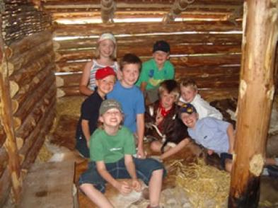 Eine Gruppe von Kindern sitzt und kniet auf Stroh in einer rustikalen Holzblockhütte, lächelt und posiert für die Kamera.