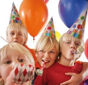 Vier Kinder mit Partyhüten und Krachmachern stehen dicht beieinander und lächeln, während im Hintergrund bunte Luftballons zu sehen sind.