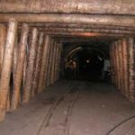 Holzbalken säumen die Wände und die Decke eines unterirdischen Bergwerkstunnels mit Gleisen am Boden.